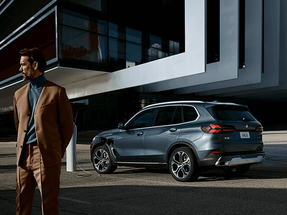 025 BMW X5 Plug-In Hybrid SUV charging outside a modern building, blending luxury and electrified performance — available now at BMW of Northwest Arkansas.