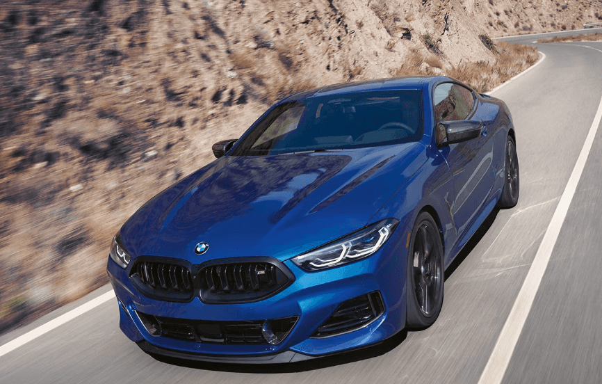 Blue 2026 BMW 8 Series coupe driving through a winding mountain road, showcasing performance and luxury at BMW of Northwest Arkansas.