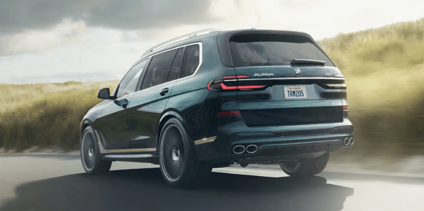 Rear view of a BMW Alpina XB7 driving through scenic countryside roads, showcasing its quad exhaust and sleek design.