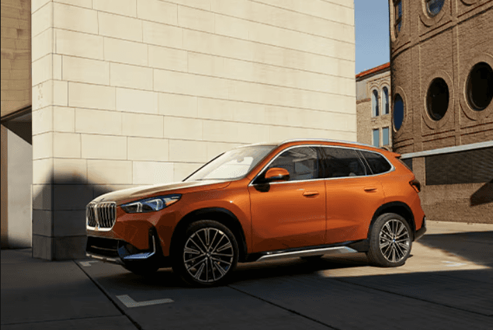2026 BMW X1 in metallic orange parked near modern buildings at BMW of Northwest Arkansas