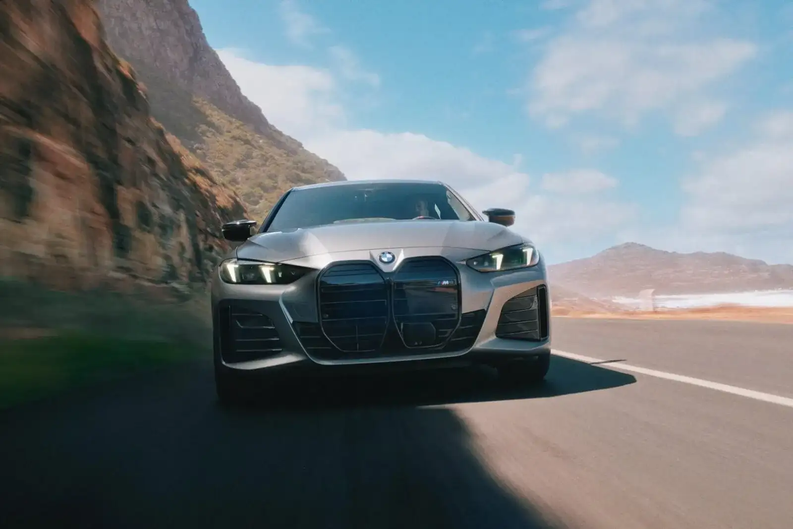 A 2026 BMW i4 M60 in motion on a coastal road, with cliffs to the left and an open shoreline to the right.