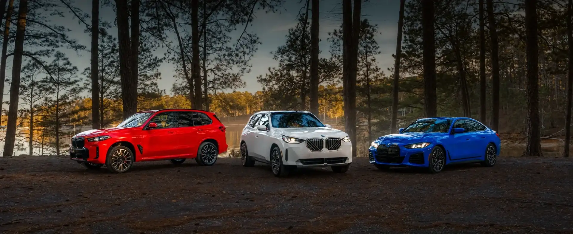 Three 2026 BMWs photographed at golden hour in a Georgia pine clearing — a red Sport Activity Vehicle on the left, a white Sport Activity Vehicle at center, and a blue sport sedan on the right, the lineup BMW of South Atlanta carries new and Certified Pre-Owned.