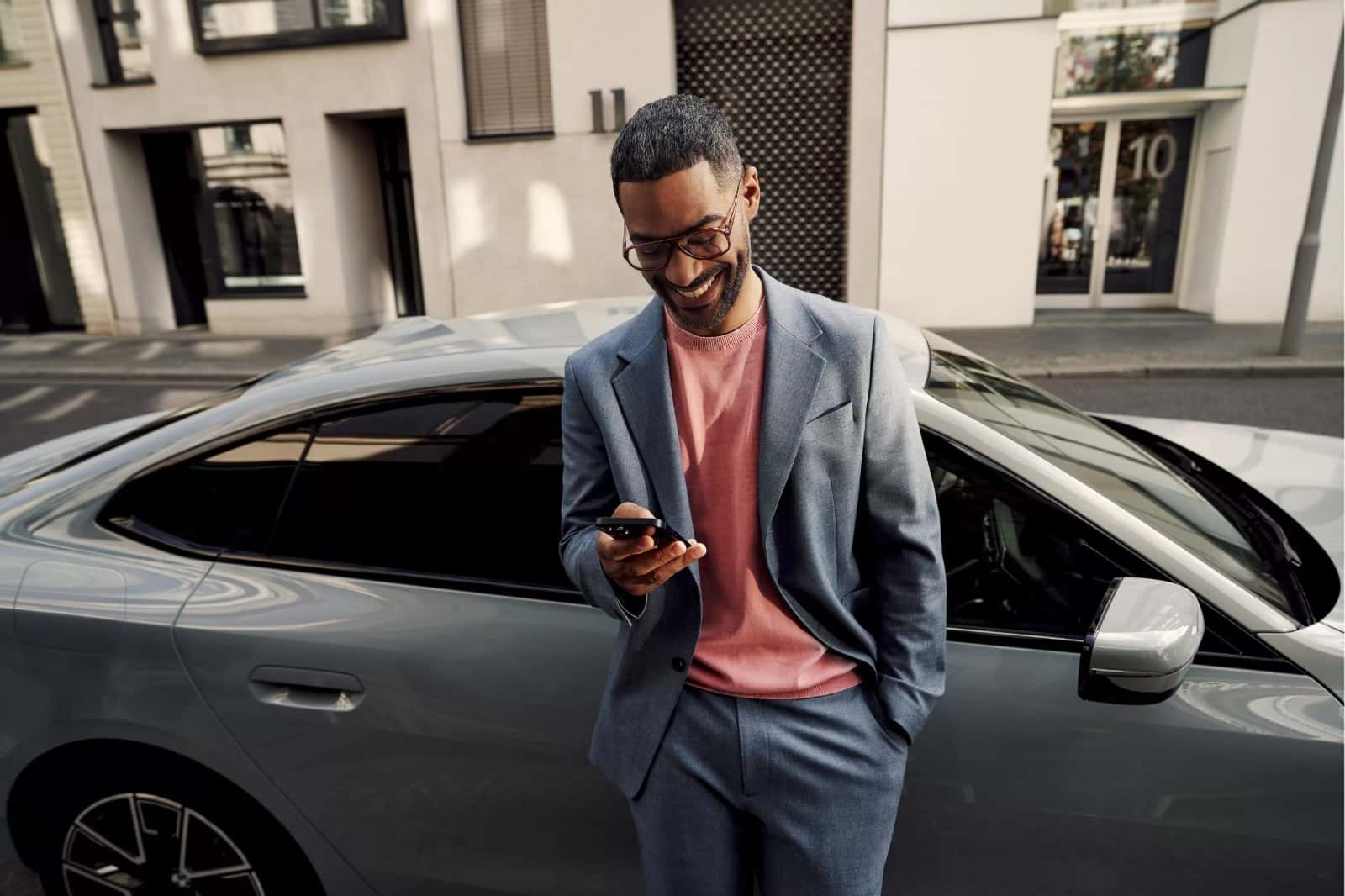 A BMW owner smiles at his phone beside his grey BMW sedan — the car is handled, the day keeps moving