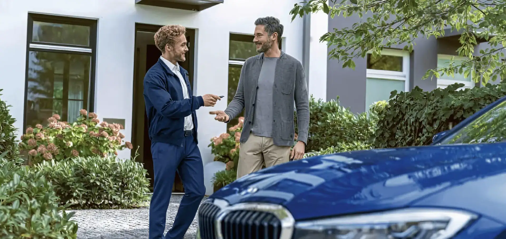 BMW service representative handing car keys to a customer in front of a home with a blue BMW in the foreground.