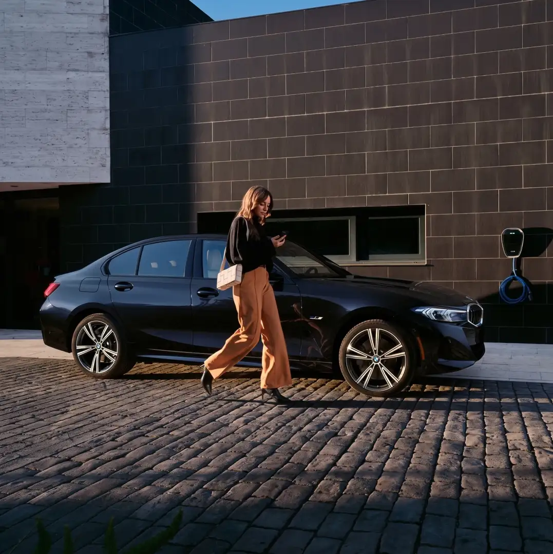 A black BMW 3 Series sedan parked on cobblestones at golden hour, a woman walking past it