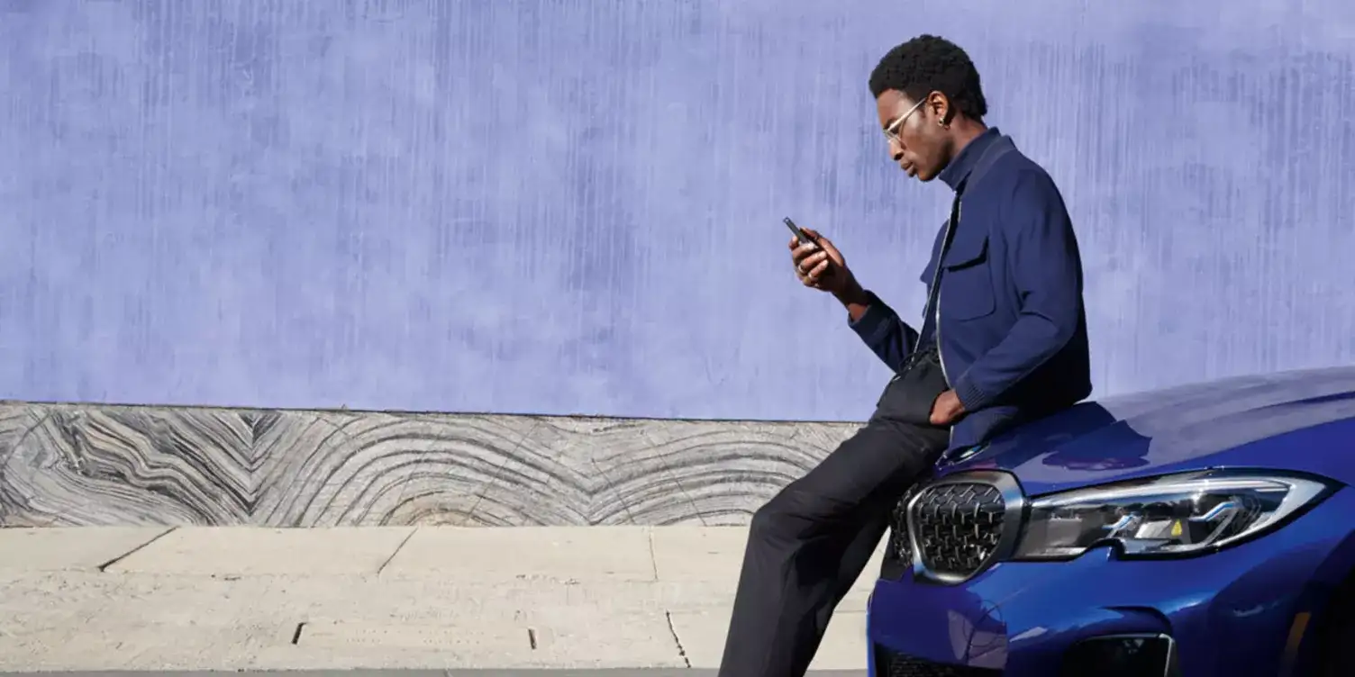 A man leaning against a blue BMW, using the My BMW app on his phone