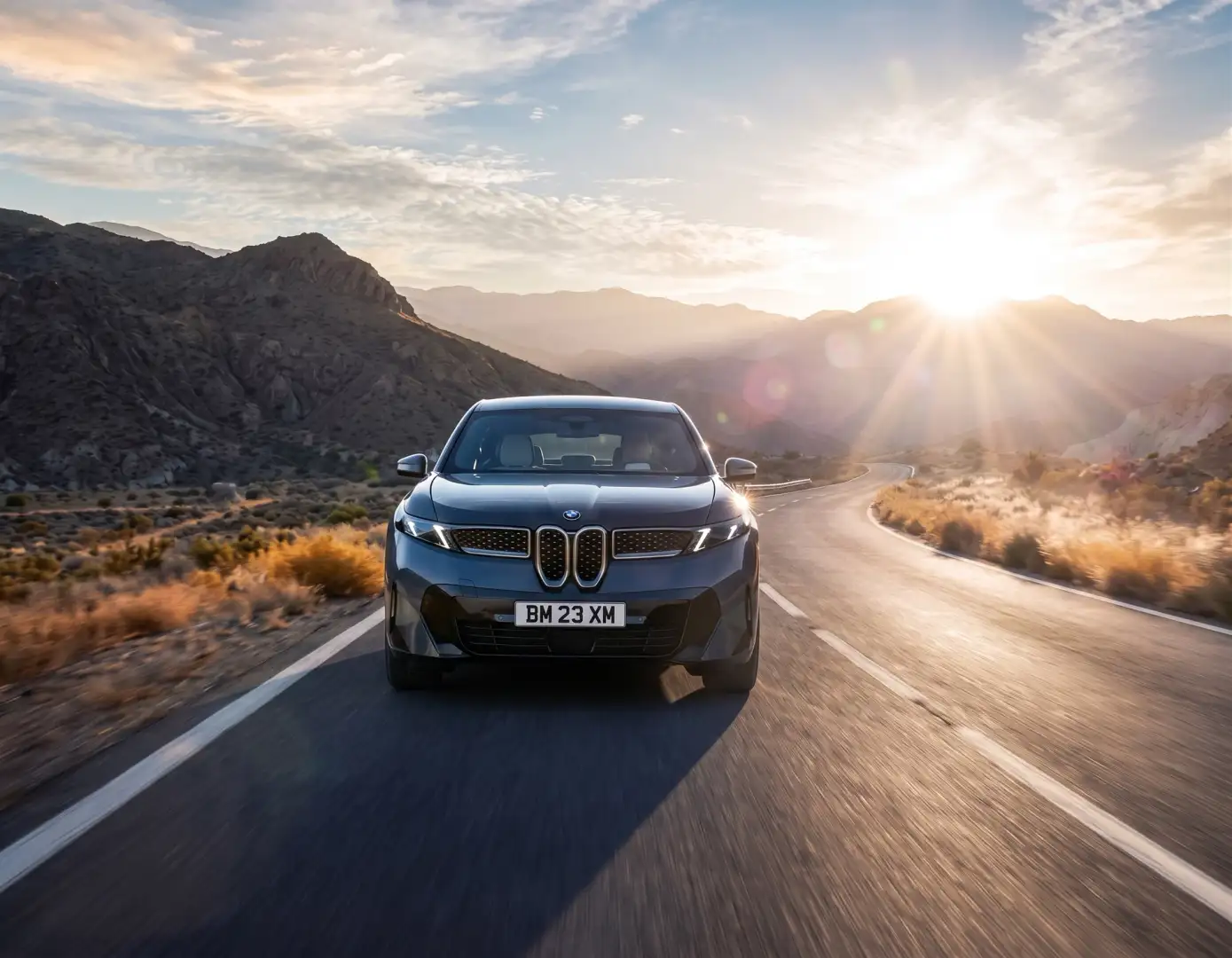 A BMW SUV driving toward the camera on a mountain road at sunrise, the sun low behind the ridge