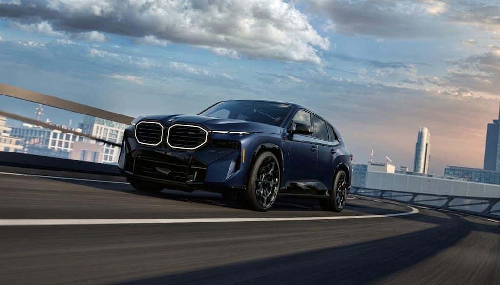 A dark blue BMW SUV driving on an elevated city road at dusk, the skyline behind it