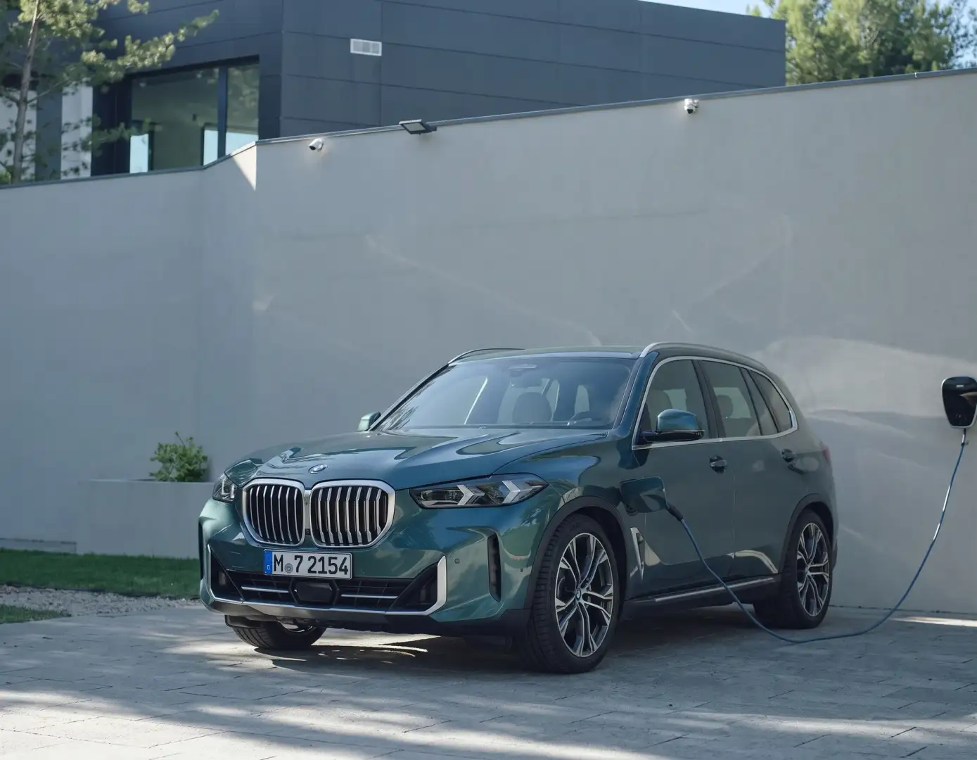 A green BMW X5 plugged into a wallbox charger on a home driveway