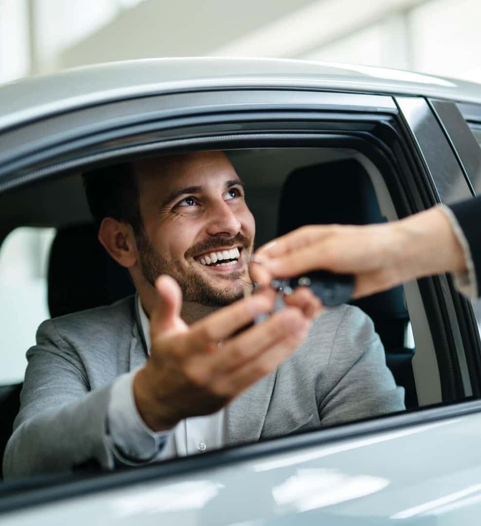 A smiling man receives car keys through the open window of his car.