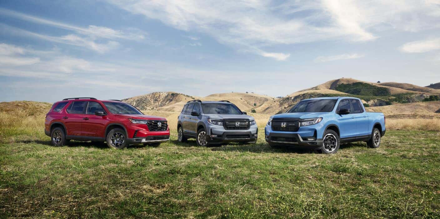 Front three-quarter view of red, gray, and blue Honda TrailSport models parked in a grassy field with hills.
