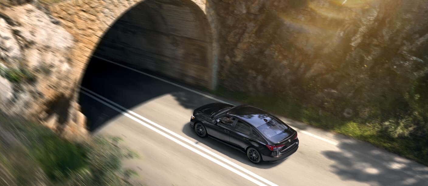 High-angle rear three-quarter view of black Honda Civic Sedan exiting a stone arch tunnel on a road.