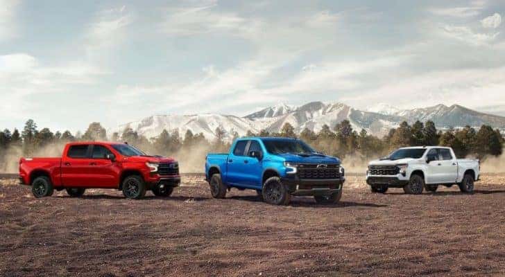 A red, a blue, and a white 2024 Chevy Silverado 1500 for sale parked on a dirt clearing.