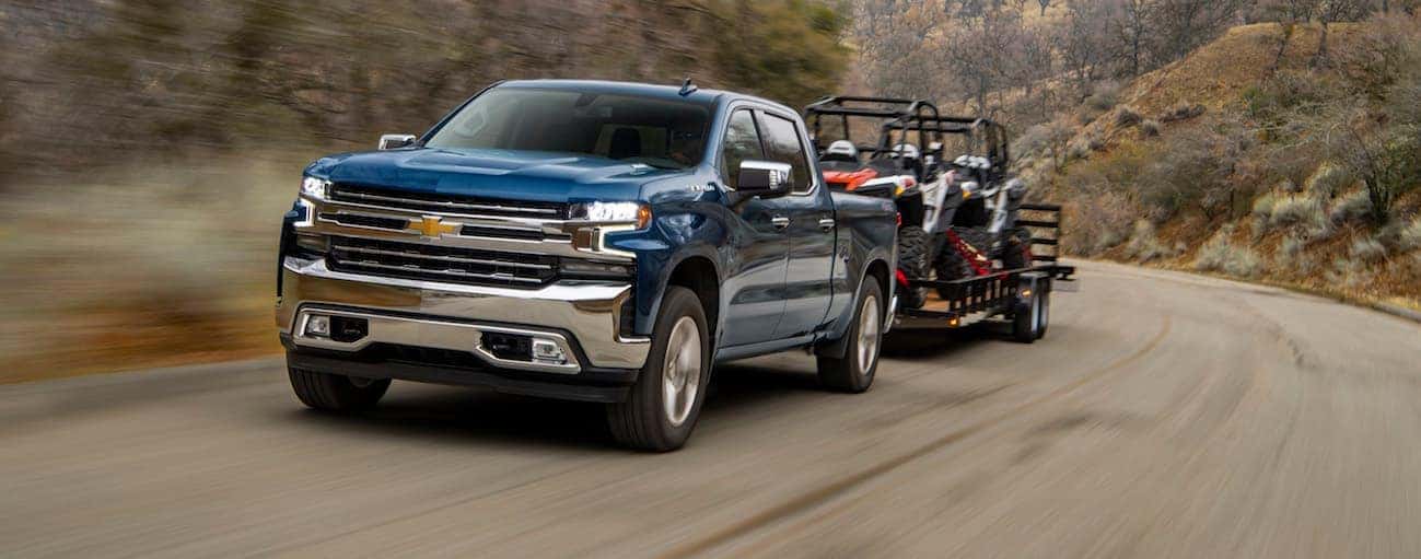 A blue 2020 Chevy Silverado 1500 is towing a trailer with side by sides on it on a dirt road.