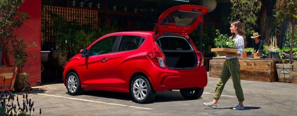 A woman is carrying a plant to the open trunk of a red 2021 Chevy Spark.