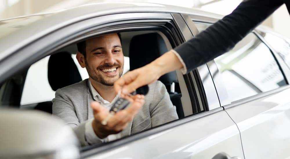 A salesman is shown handing a man car keys while he sits in a silver sedan.