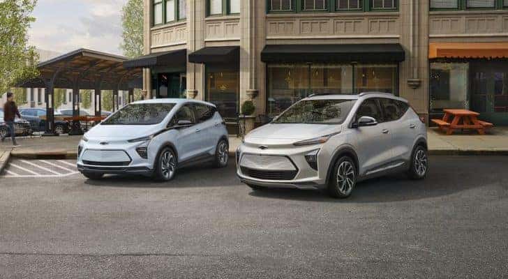 A light blue 2023 Chevy Bolt EV and silver EUV are shown parked in front of a shop.