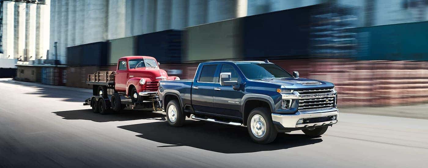 A blue 2022 Chevy Silverado 2500HD is shown towing a red truck on a city street.