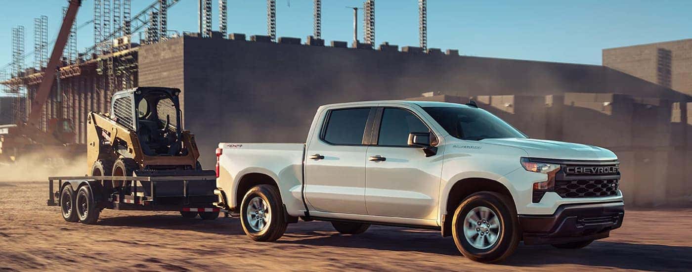 A white 2023 Chevy Silverado 1500 WT is shown towing heavy machinery on a trailer.