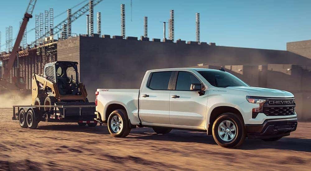 A white 2023 Chevy Sivlerado Custom is shown from the side while towing a skid-steer.