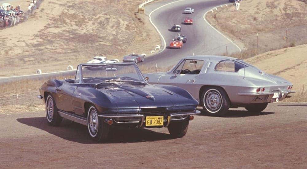 Two 1963 Chevy Corvettes are parked with a racetrack behind them.