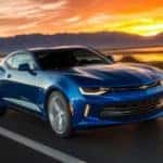 A blue 2016 Chevy Camaro is driving on a highway in front of a vibrant sunset.