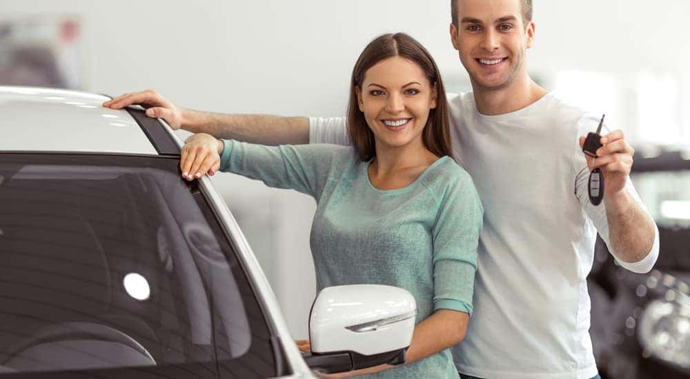 A couple is leaning on a white car with the keys in the man's hand.