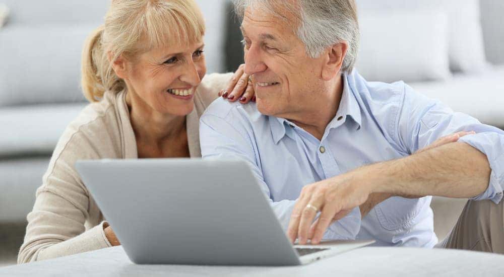 An elderly couple is smiling while using a laptop in their living room.