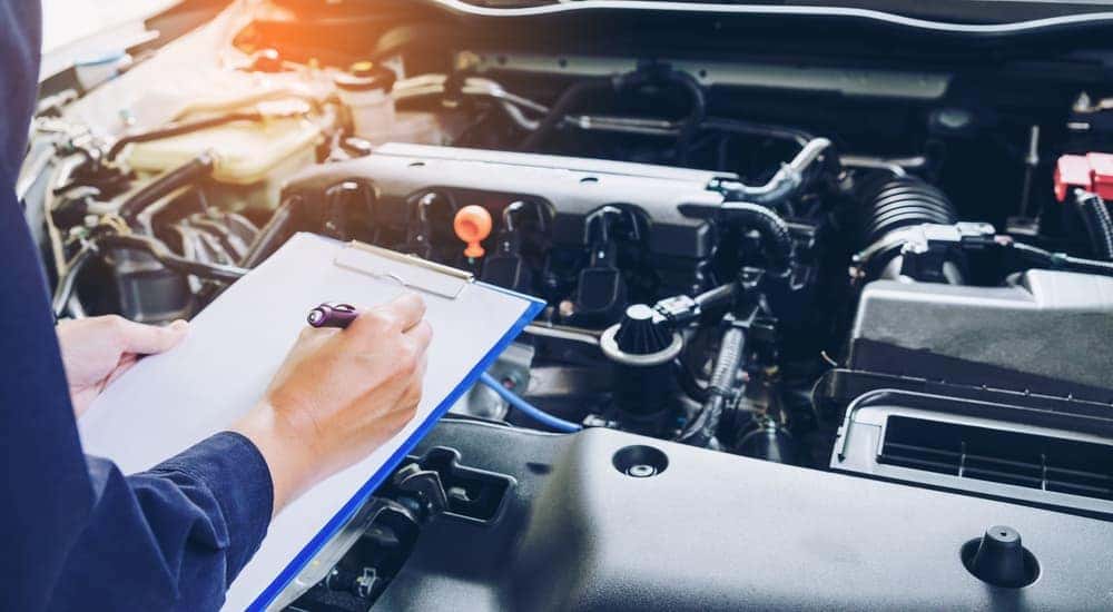 A mechanic is using a clipboard while looking at the motor of a car.