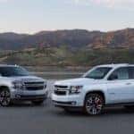 The largest of the Chevy SUV models, a silver 2020 Chevy Suburban and a white 2020 Chevy Tahoe, are parked in front of a lake and mountains.
