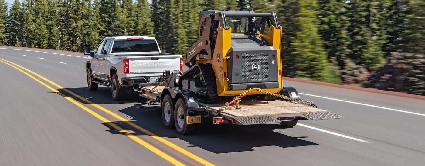 A white 2020 Used Chevy Silverado 2500HD is towing construction equipment past pine trees.