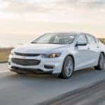 A white 2018 Chevy Malibu is driving along a coastal highway.