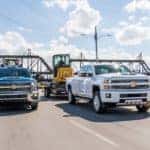 A white and a blue 2018 Chevy Silverado 2500HD are towing construction equipment past a bridge on a highway.