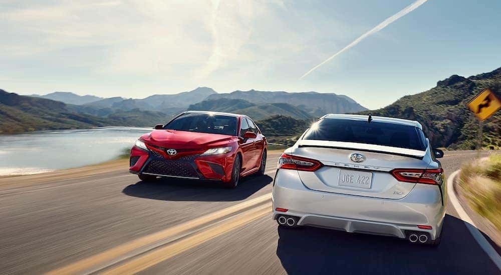 A white and a red 2018 Toyota CAmry are passing each other on a road past mountains and lakes.