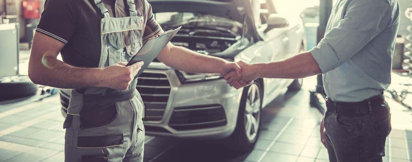 A mechanic is shaking hands with a car owner in front of a white car with the hood open.