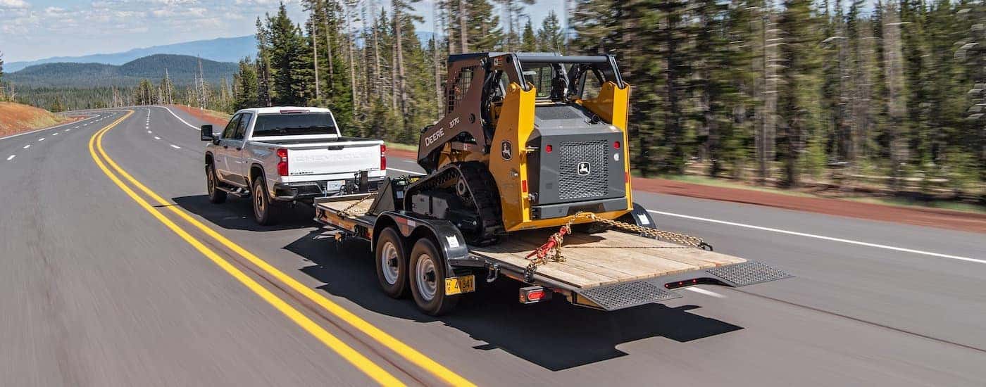 A white 2021 Chevy Silverado 2500 HD is shown from behind towing construction equipment down a tree-lined highway.