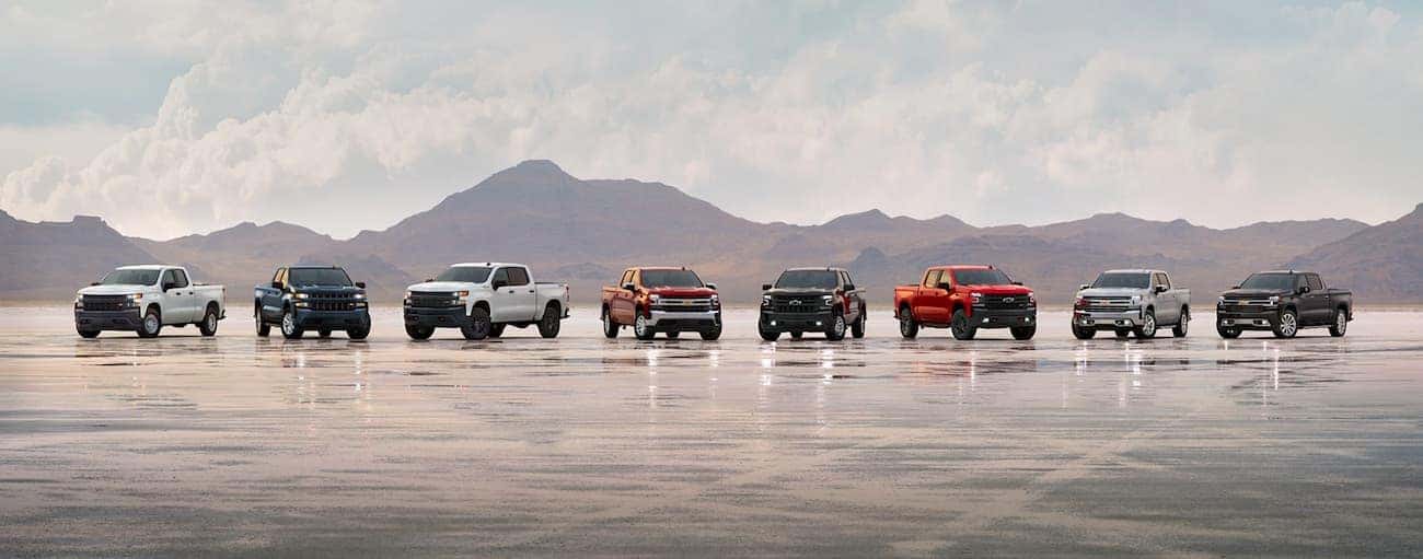 Eight 2019 Chevy Silverado 1500s are parked in a line on a salt flat in front of mountains.
