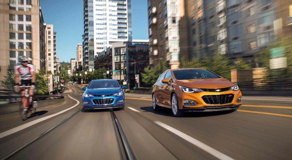 An orange 2018 Chevy Cruze Hatchback RS is driving next to a blue Cruze in a city.