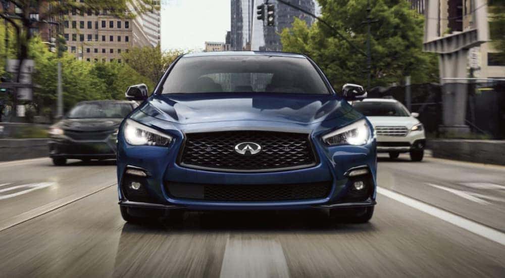 A blue 2021 Infiniti Q50 is shown from the front driving on a city road after searching 'pre-owned luxury car sales'.