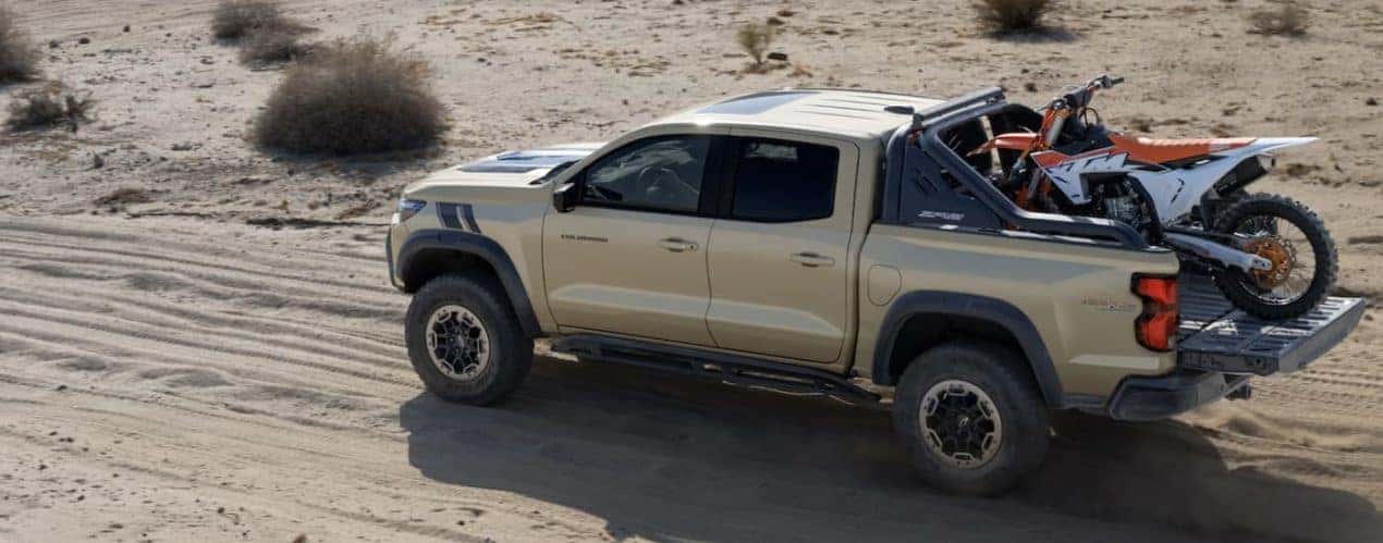 A tan 2023 Chevy Colorado Trail Boss is shown driving with a dirt bike in the bed.