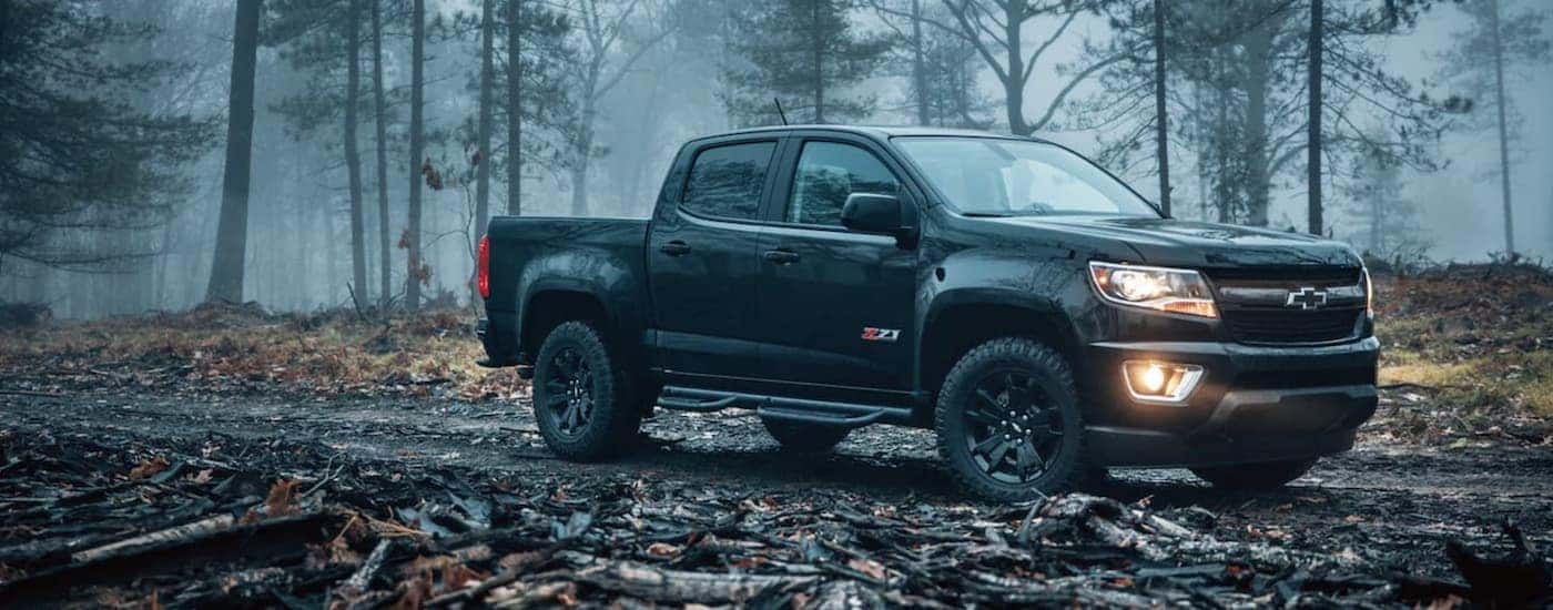 A popular used truck for sale, a black 2018 Chevy Colorado Z71, is shown from the side parked in the woods.