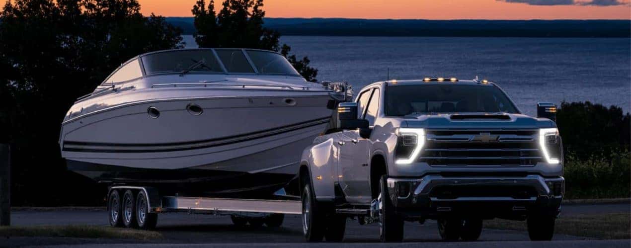 A white 2024 Chevy Silverado 3500 HD is shown towing a white boat during a sunset.