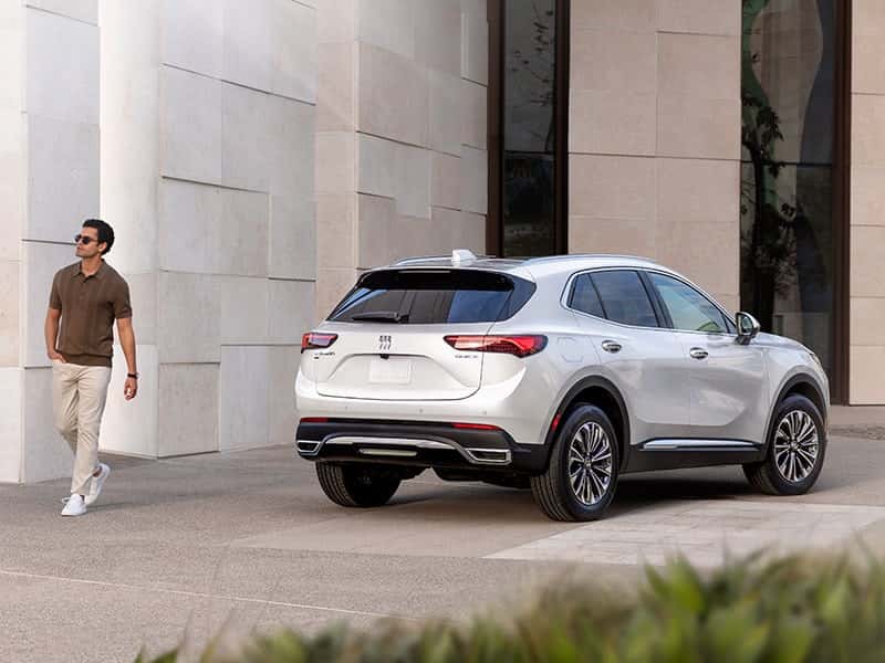 A rear view of a silver Buick Envision, parked by a building as a man wearing sunglasses walks away from it