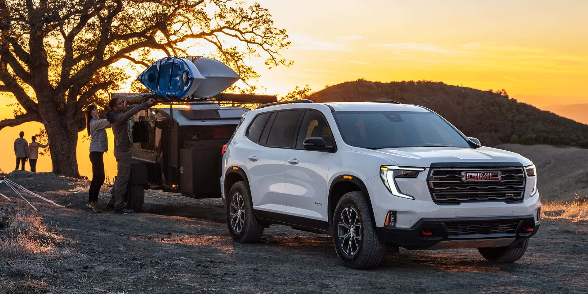A white GMC Acadia connected to a trailer with a family loading the trailer