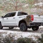 A white 2022 Chevy Silverado 1500 Realtree Edition is shown from the rear at an angle parked in a snowy forest after leaving a Chevy Dealer in Minot.