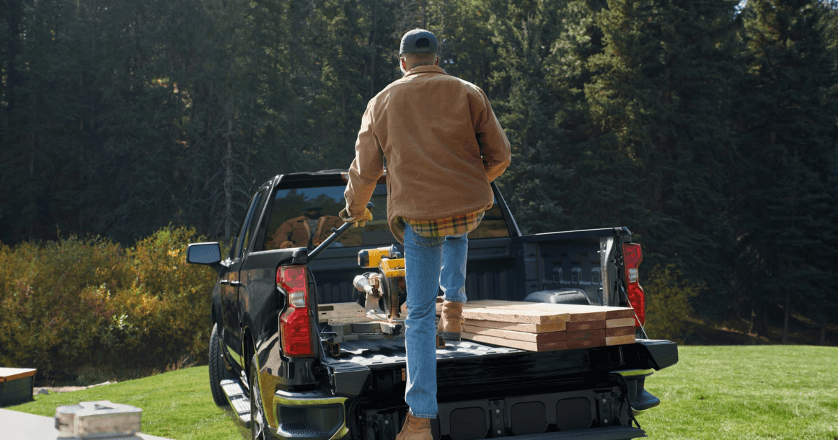 A black Chevrolet Silverado 1500 with a man climbing into its truck bed with a saw and lumber