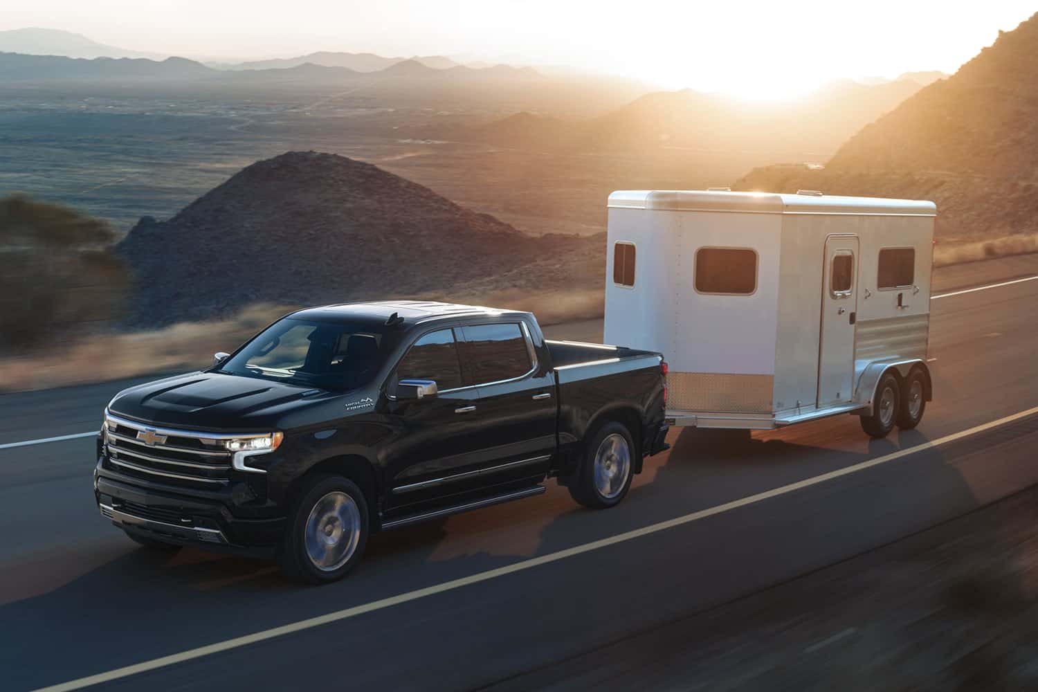A black Chevy Silverado 1500 driving down the road in a hilly region and towing a small trailer
