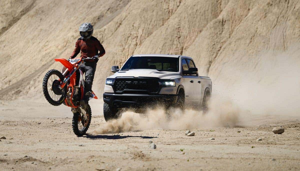 A white RAM 1500 driving on dirt behind a dirt bike