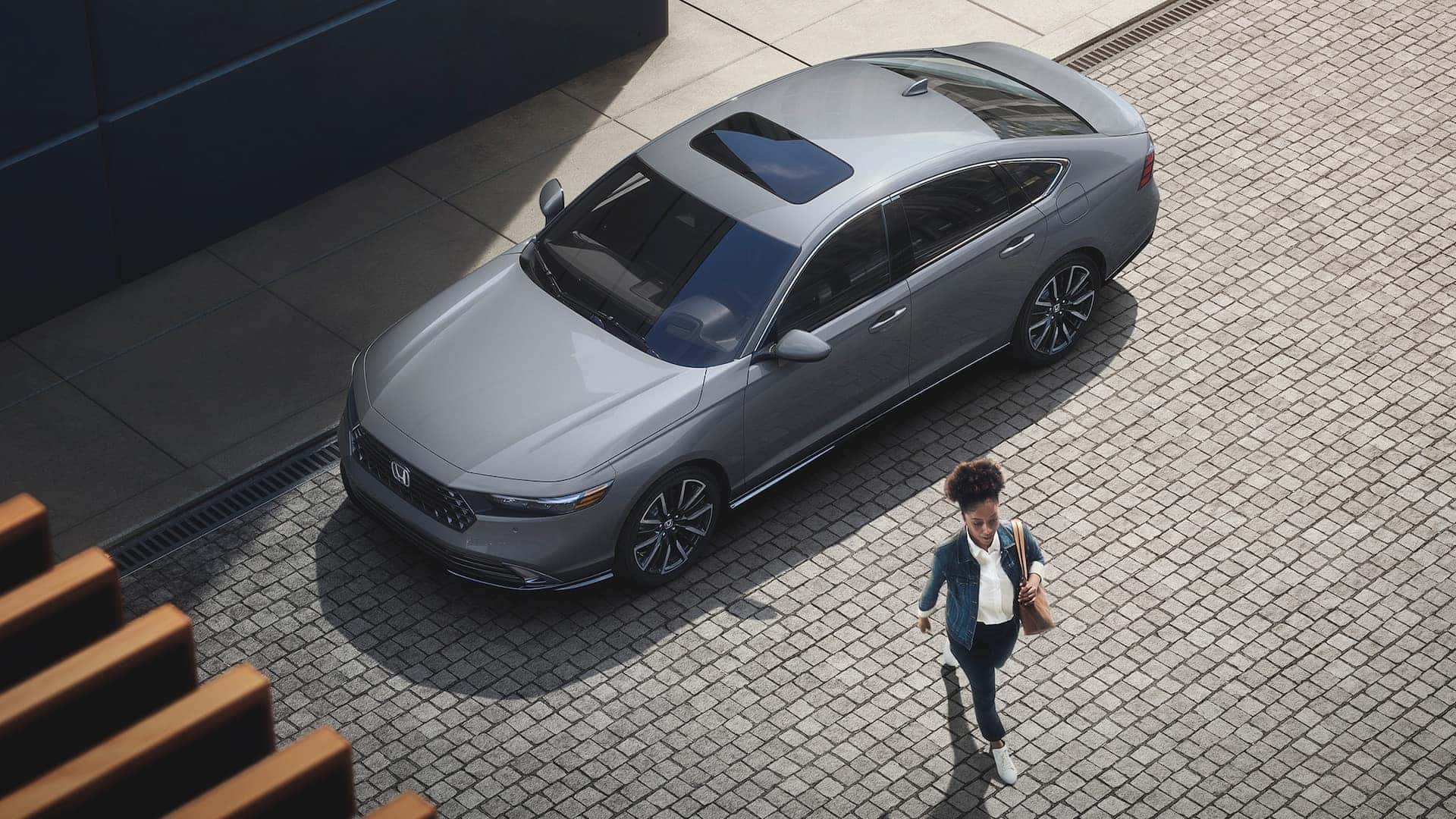 A gray 2025 Honda Accord hybrid parked by a sidewalk with a woman walking away from it