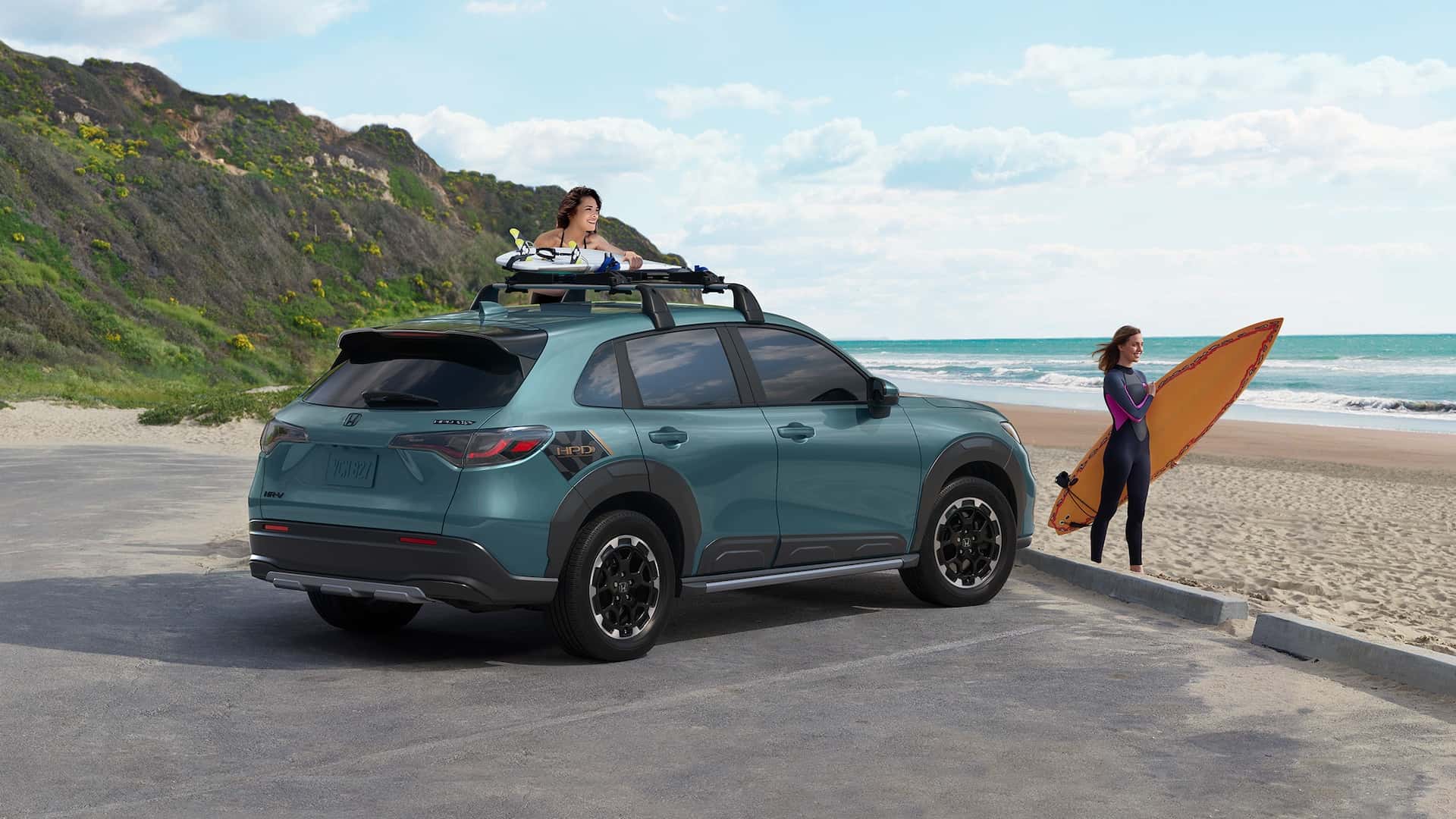 A turquoise 2025 Honda HR-V parked by a beach with a woman getting their surfboard off the top while another woman walks in front with her surfboard.